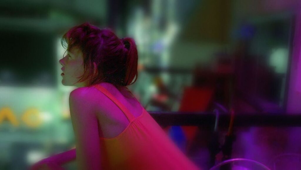 Enter the Void — Gaspar Noe Woman in neon lighting. Back view of a woman with short hair in a pink top, looking to the side in a blurred, colorful setting.