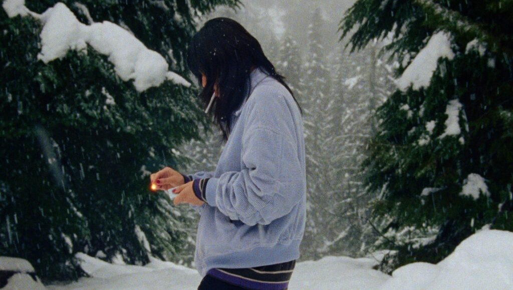 Prismatic Ground 2022 — Dispatch 2: Lake Forest Park, Acts of Love, Color Without Colour Woman lights match in snowy forest. Winter landscape with snow-covered trees. Cold weather scene.