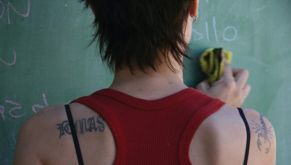 Lion’s Den — Pablo Trapero Woman erasing chalkboard. Tattoos on shoulders: text and scorpion. Red tank top. Education concept.