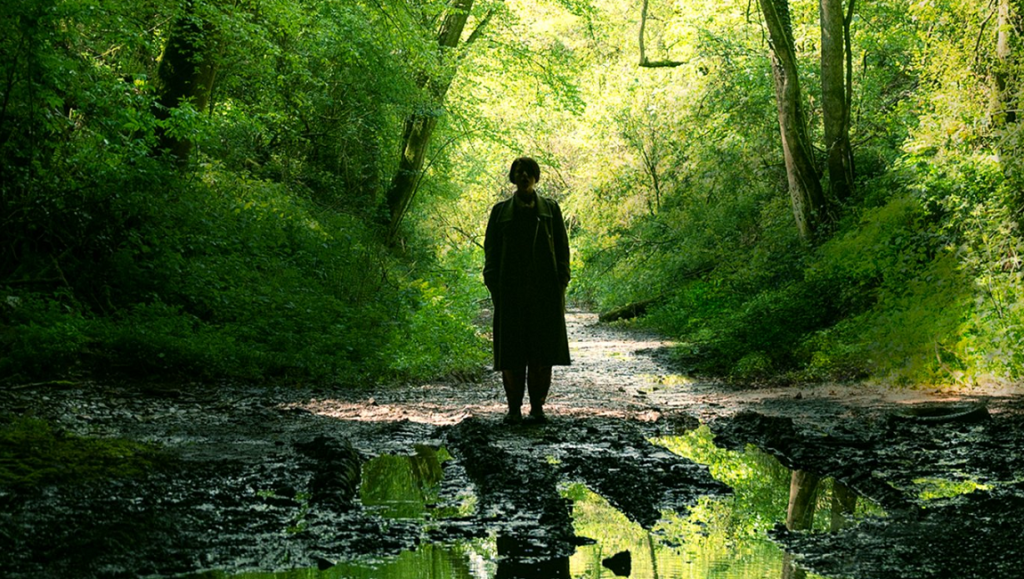 Men movie image. Woman standing on a muddy path in a green forest. Alex Garland film.