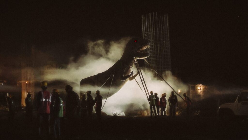 Once Upon a Time in Calcutta movie scene: A large dinosaur prop being set up at night, surrounded by crew.