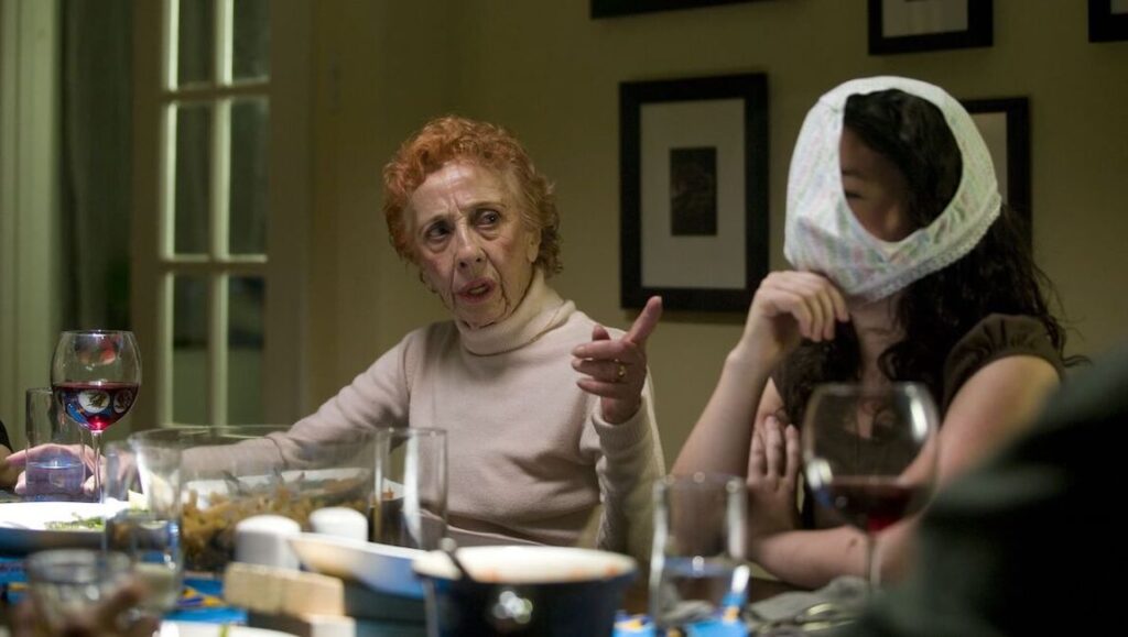 Elderly woman gesturing at a dinner table, next to a woman with underwear on her head. Comedy scene.