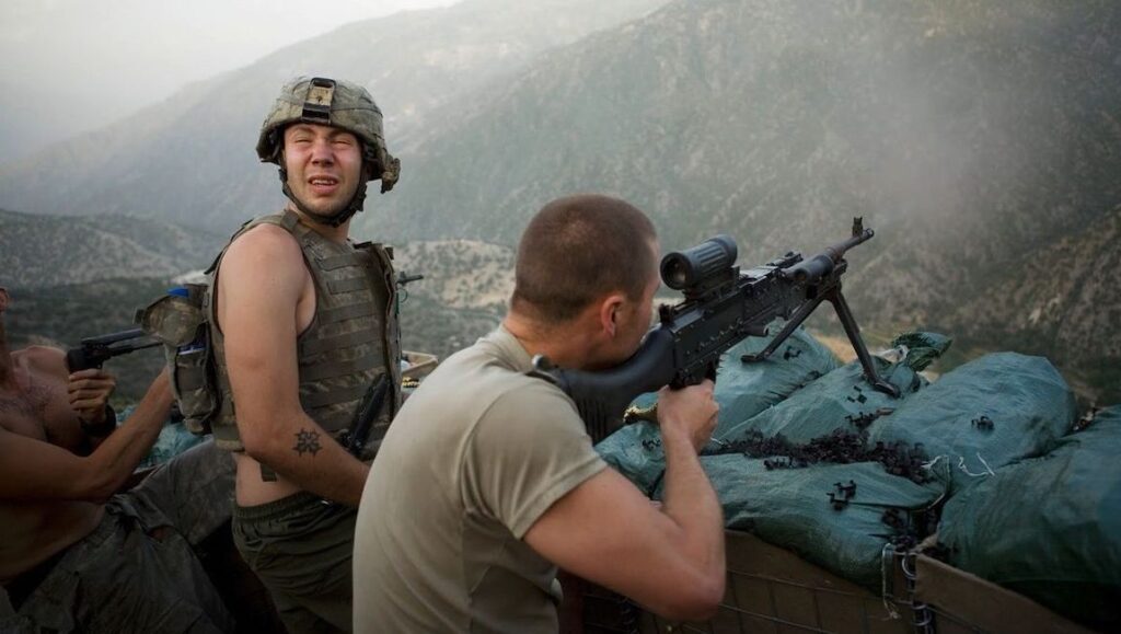 Restrepo — Tim Hetherington and Sebastian Junger Soldiers in combat. One aims a machine gun from a sandbag bunker. Another soldier reacts to gunfire. Mountain backdrop.