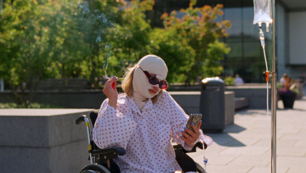 Sick of Myself film still. Woman in bandages, sunglasses, and hospital gown in a wheelchair, smoking and using a phone.
