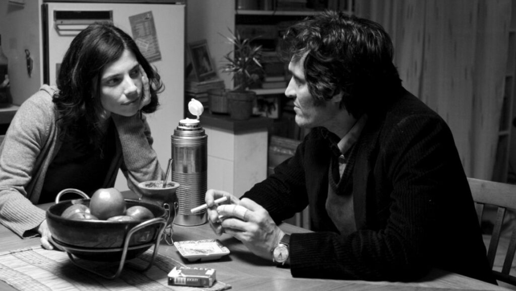 Black and white film still. Man smokes, woman listens. Mate gourd, thermos, fruit bowl on table. Drama, conversation.