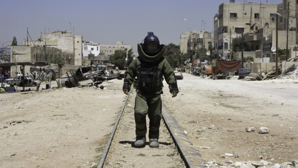 The Hurt Locker movie scene: bomb disposal expert in protective suit walks on train tracks in war-torn city.