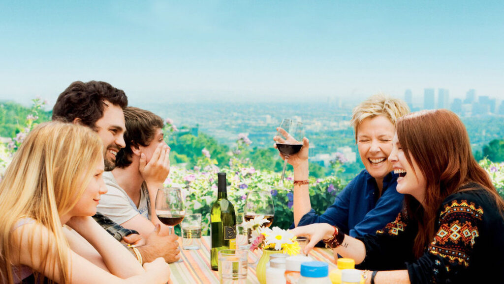 The Kids Are All Right movie scene. Family laughs at outdoor table with wine, city view. Annette Bening, Julianne Moore.