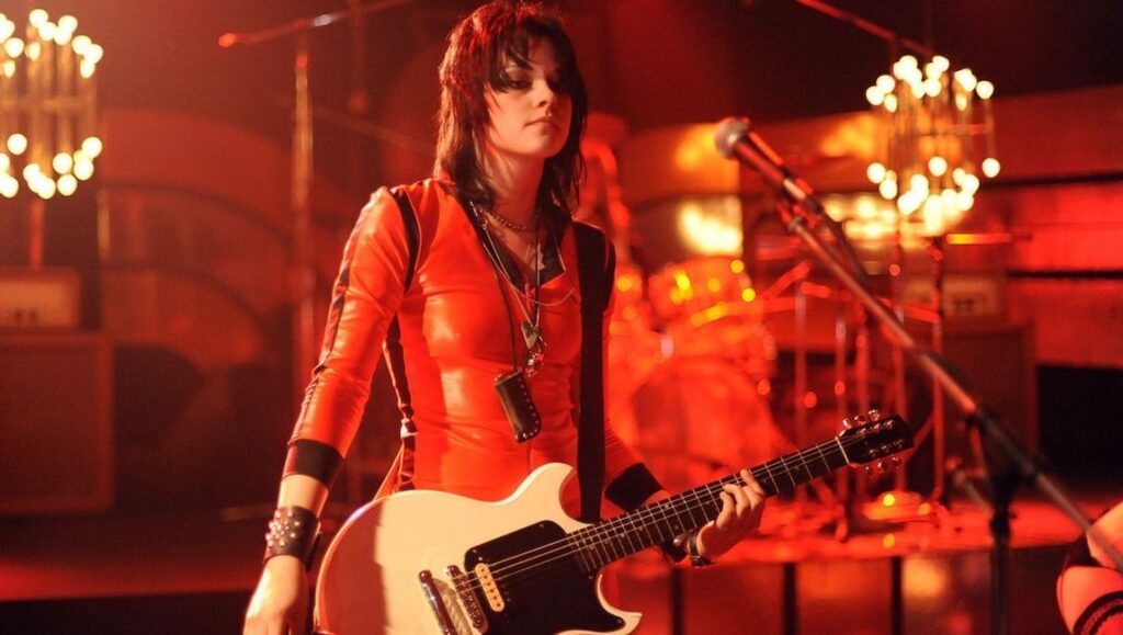 Kristen Stewart as Joan Jett in The Runaways movie. Rock star playing guitar on stage. Red lighting.