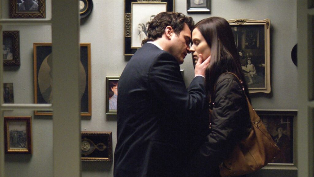 Two Lovers movie still. Joaquin Phoenix and Gwyneth Paltrow embrace in front of a wall of framed photos.