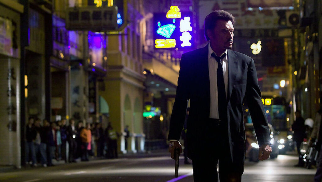 Vengeance — Johnnie To Man in suit with gun walking on city street at night. Vengeance movie scene.