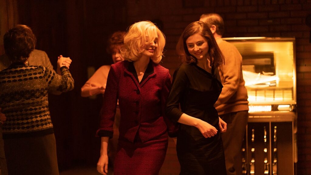 Eileen film scene: Anne Hathaway and Thomasin McKenzie at a party with a jukebox.