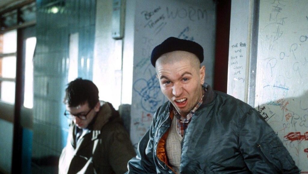 Meantime film still. Tim Roth as skinhead, aggressive expression, beret. Graffiti background. 1980s British cinema.