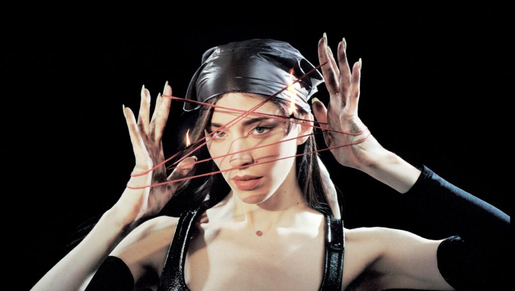 Caroline Polachek portrait. Woman with red strings, headscarf, and long nails against a black background. Music artist.