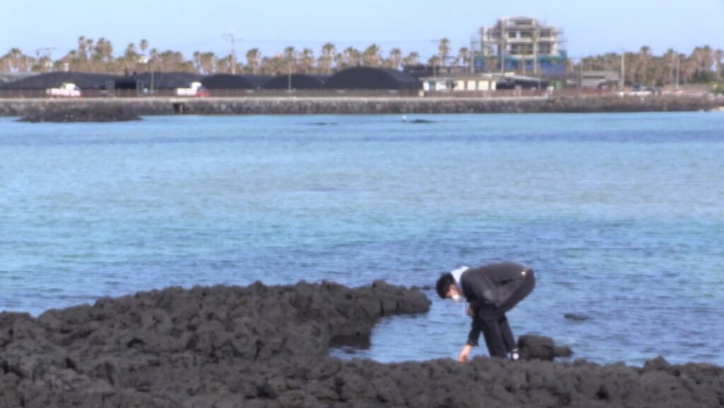 Jeonwonsa Film Co. in water. Man in suit on rocky shore near ocean. Coastal scene with buildings in background.