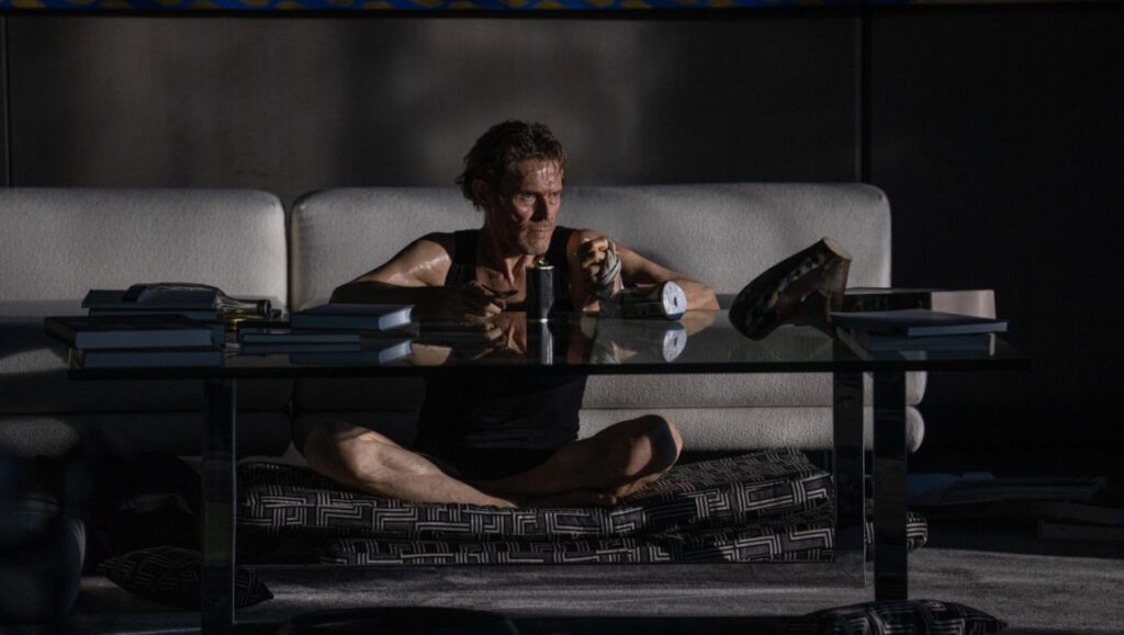 Heretic: Willem Dafoe sits cross-legged at a glass table. Books and objects are on the table. He is sweaty.