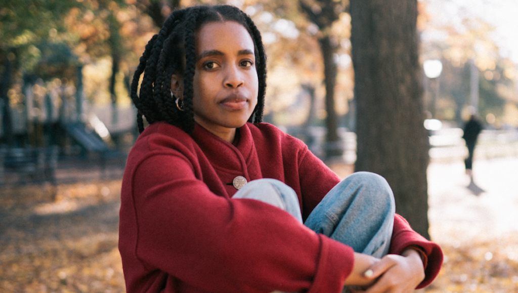 Woman with braids wearing a red coat and jeans sitting in a park. Fall foliage.