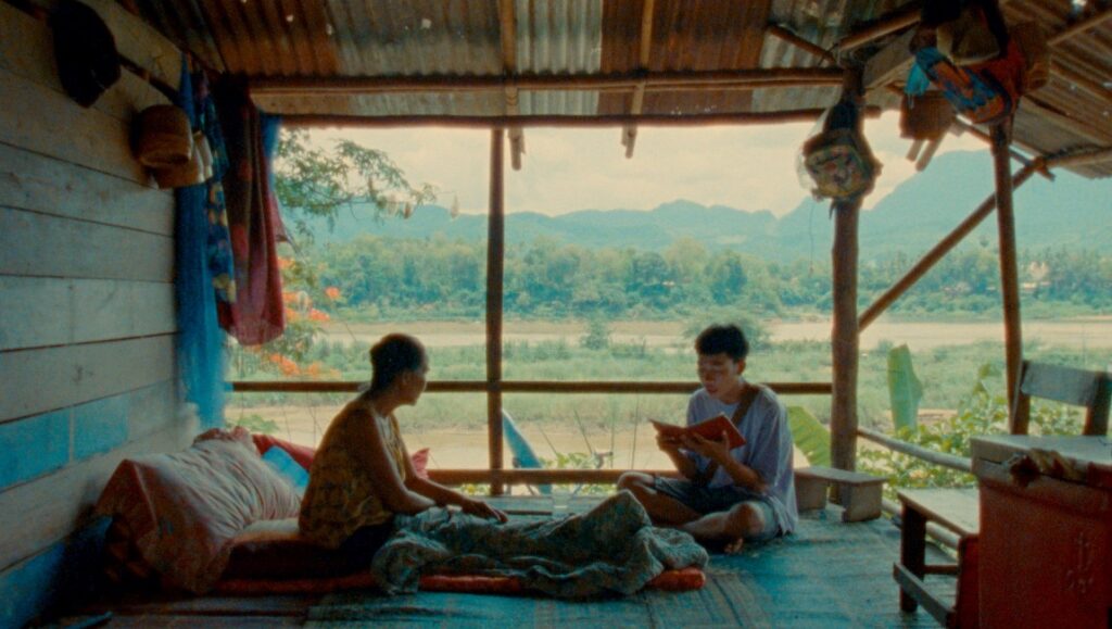 Indoor scene with two people, one reading a book, with a river and mountain view in the background.