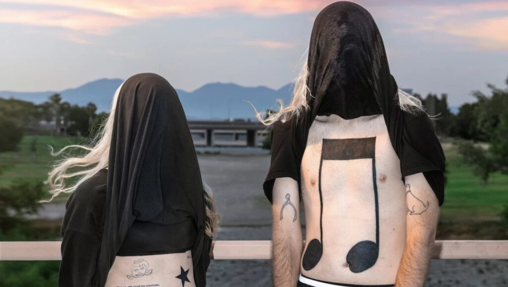 100 Gecs' 10,000 Gecs album art. Two figures with covered faces and musical note body art stand against a sunset backdrop.