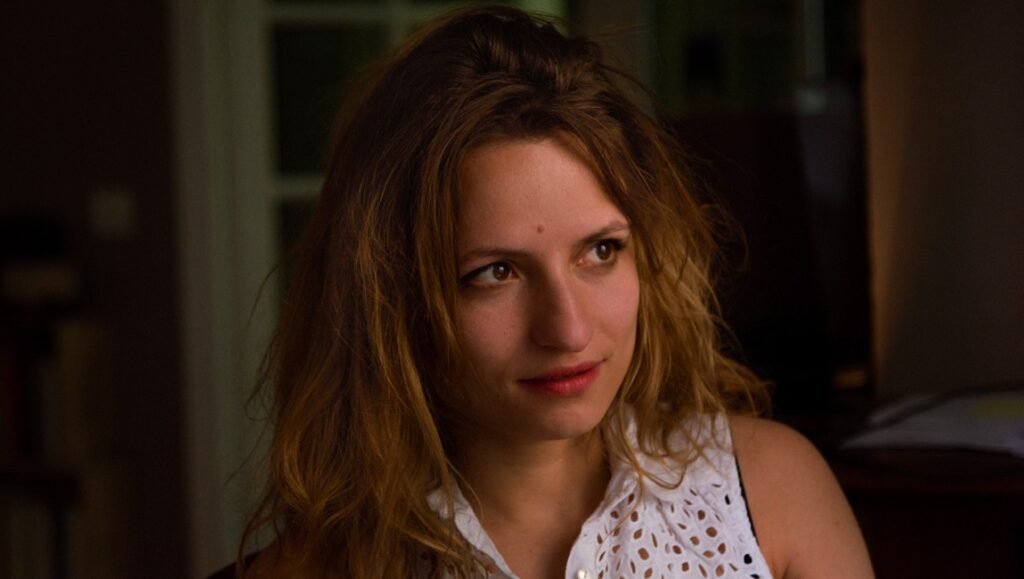 Headshot of a female director with wavy brown hair, wearing a white top. Portrait by Paul Guilhaume.
