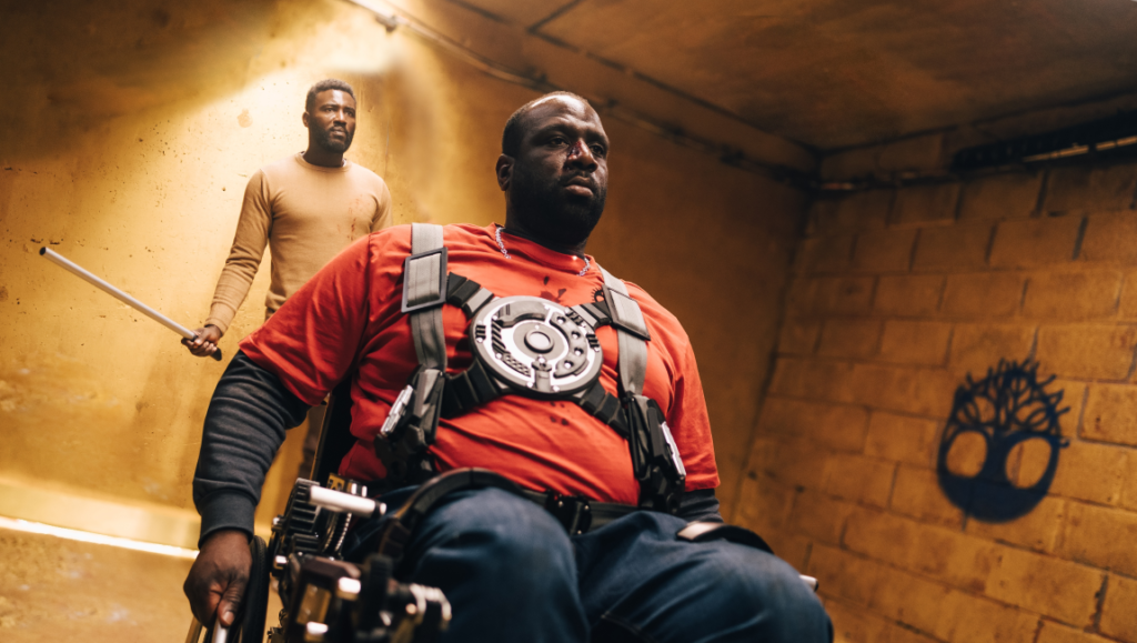 Three Nights a Week movie scene. Man in wheelchair with tactical gear, another man stands behind him holding a rod.