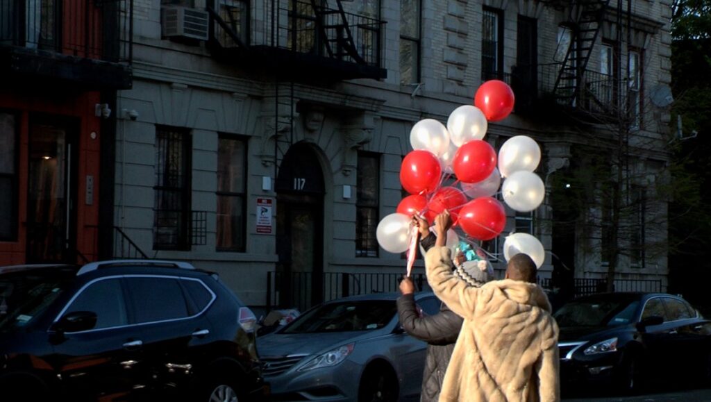 Su Friedrich's Today film still. People hold red and white balloons outside building #117. Cars parked on street.