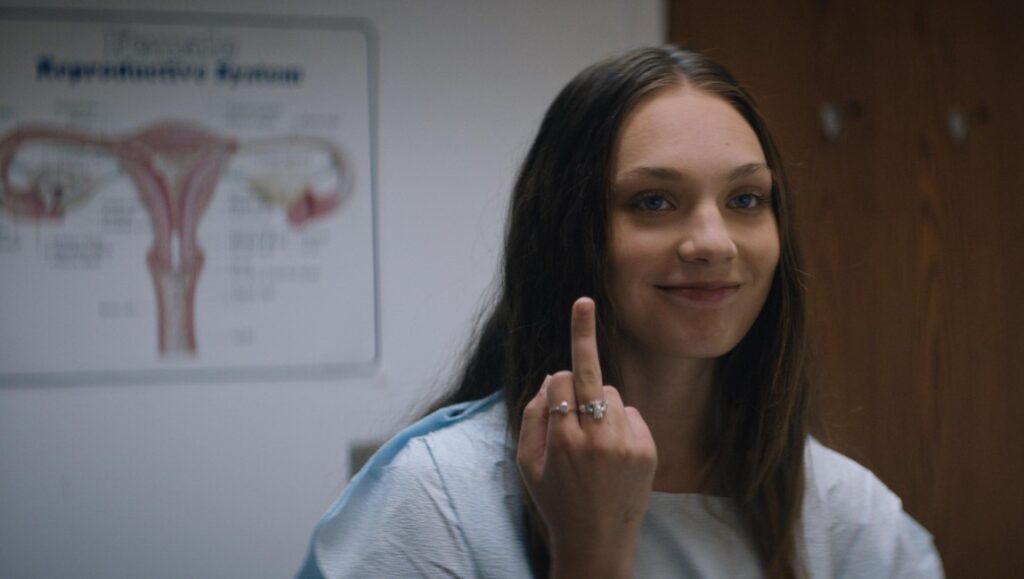 Fitting In movie scene: Woman flipping off the camera in a doctor's office, uterus diagram behind her. Film review.