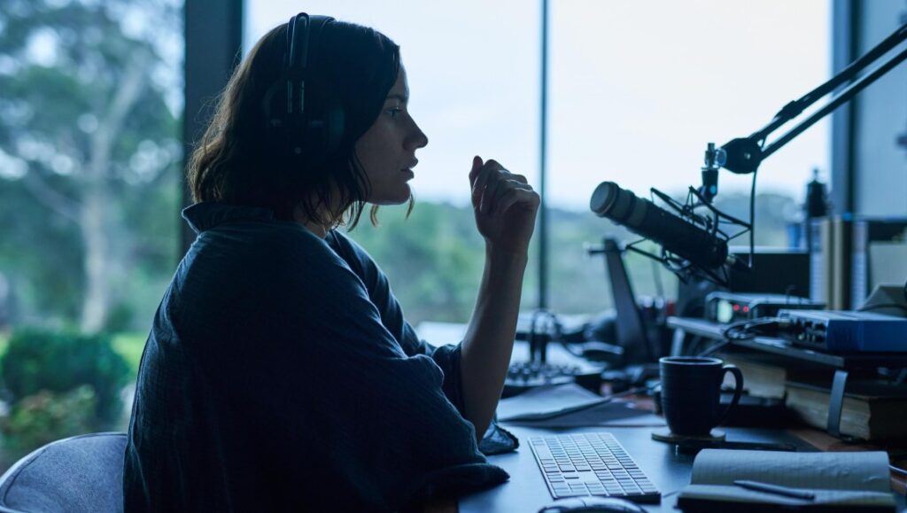 Monolith film review: Woman podcasting with headphones and microphone. Dark, moody lighting.