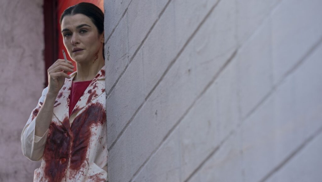Rachel Weisz in Dead Ringers. Woman in blood-stained coat smokes cigarette, leaning against wall. Prime Video series.