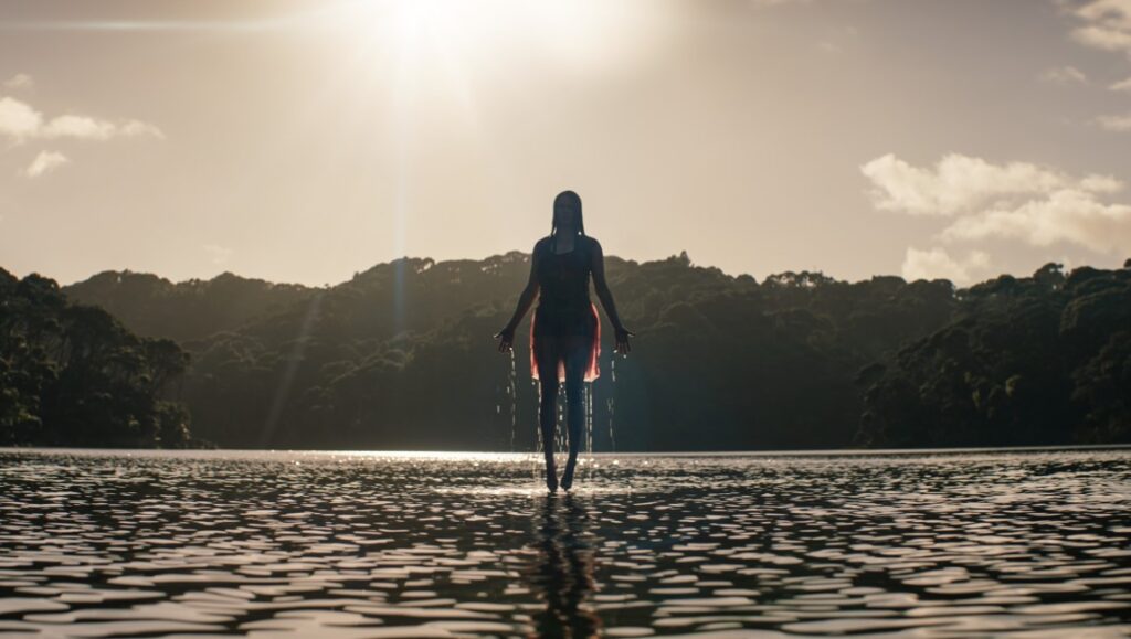 Eden: Reborn. A woman stands in the water, backlit by the sun. Lush green trees line the shore.