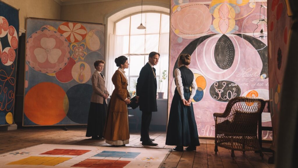 Hilma af Klint exhibition. People viewing abstract paintings with geometric shapes and floral motifs. Art gallery scene.