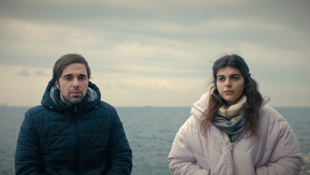 Almost Entirely film still. Man and woman in winter coats by the sea. New Directors/New Films Festival.