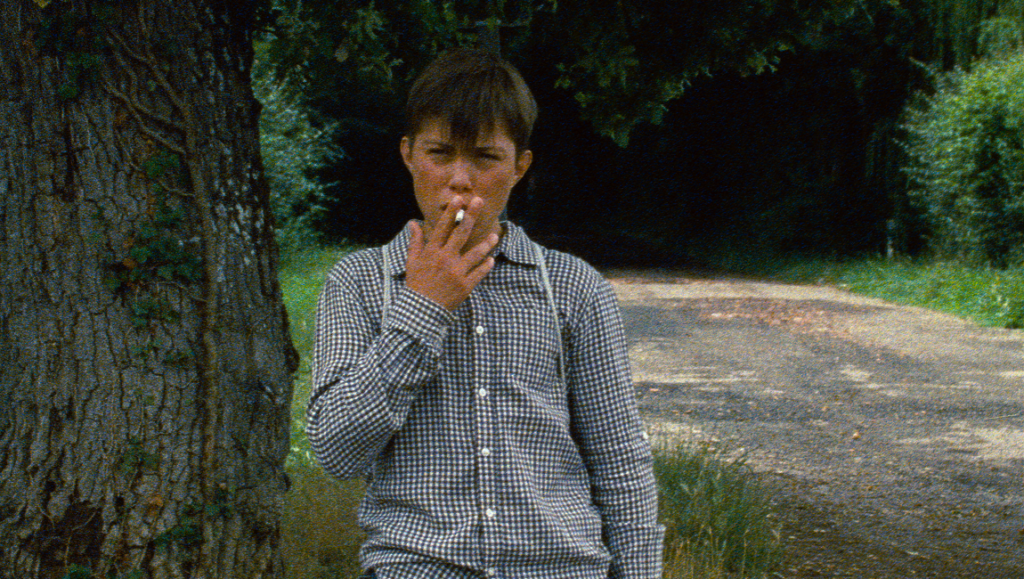 Astrakan film still: Boy smoking. Young boy in a checkered shirt and suspenders stands by a tree, holding a cigarette.