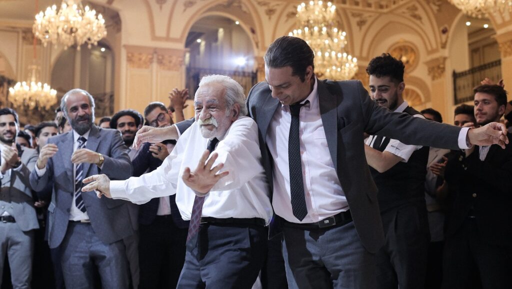 Leila's Brothers film scene: Two men dancing at a celebration, surrounded by cheering people in formal attire.
