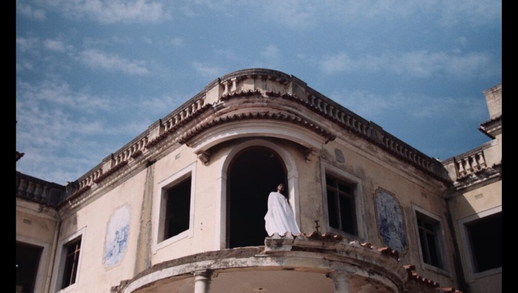Nakuzandza film still: Person in white on balcony of aged Maputo building, blue sky. Art, architecture.