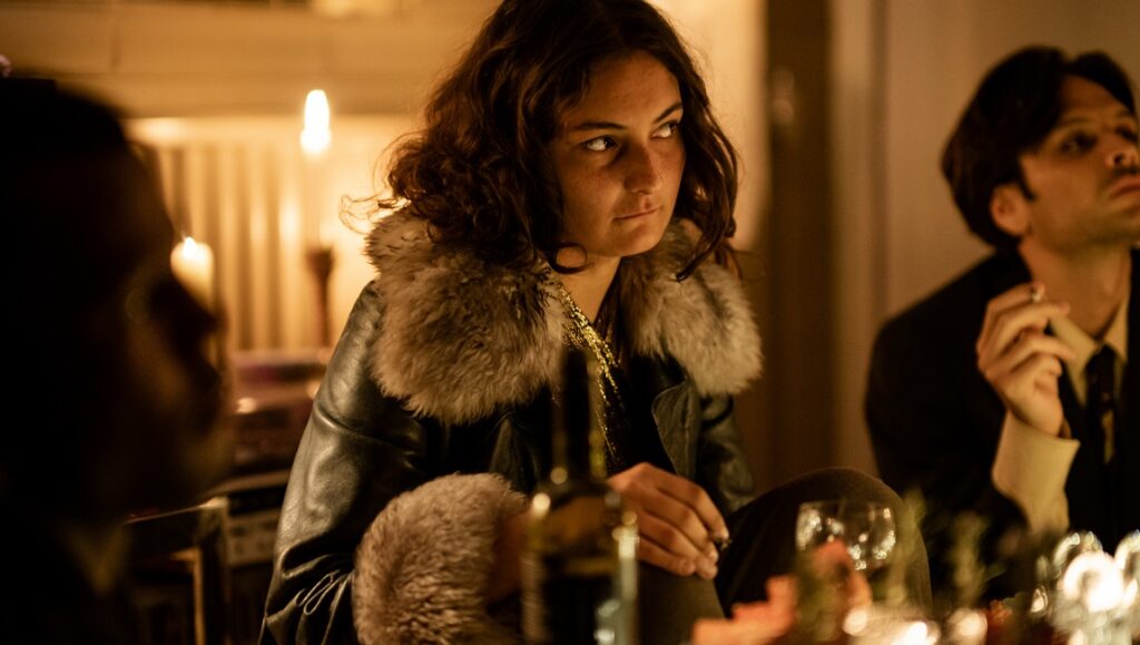 Petrol film scene: Woman in fur-collared jacket at a table with other people. NDNF 2023.