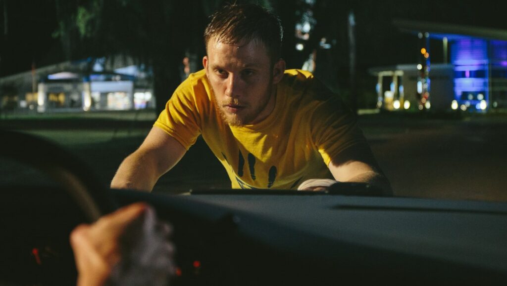 One Of These Days movie scene. Man in yellow shirt leans on a car at night, looking ahead. Beau Coulon. Flare Film.