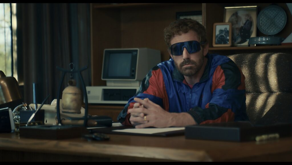 Air — Ben Affleck Ben Affleck as Phil Knight in Air movie. Knight sits at a desk wearing sunglasses and a blue and red jacket, with a vintage computer in the background.