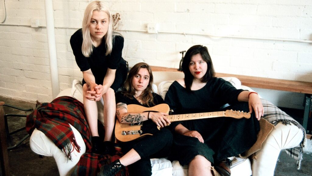 Boygenius band portrait. Phoebe Bridgers, Julien Baker, and Lucy Dacus sit on a couch with a guitar. Indie rock group.