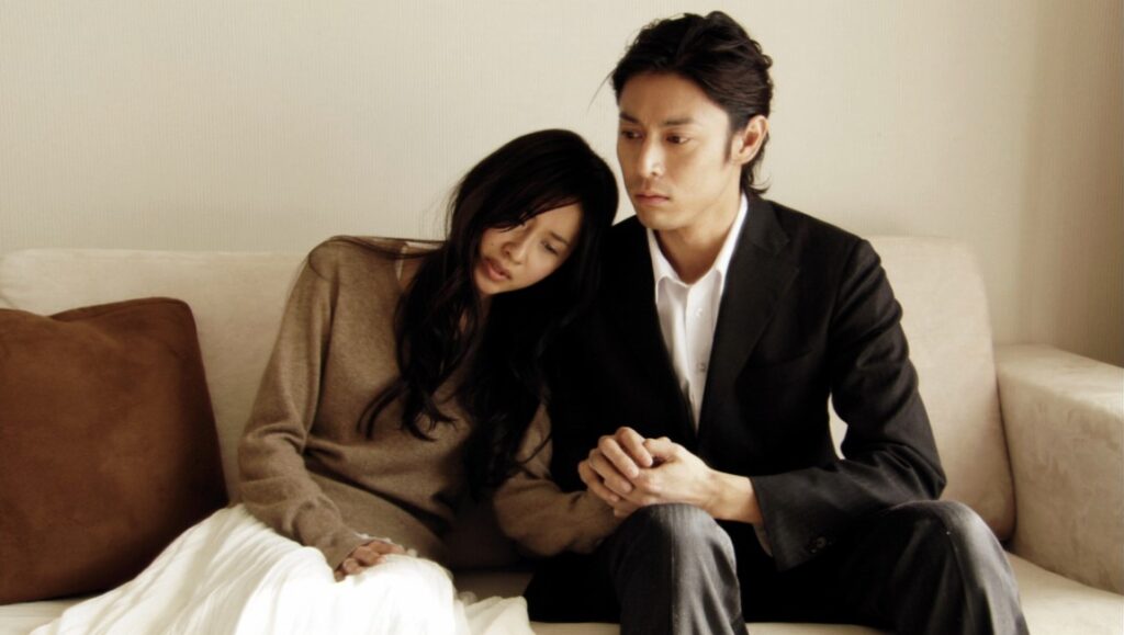 Passion — Ryûsuke Hamaguchi Film still. Couple on couch. Woman rests head on man's shoulder. He wears a suit. Drama.