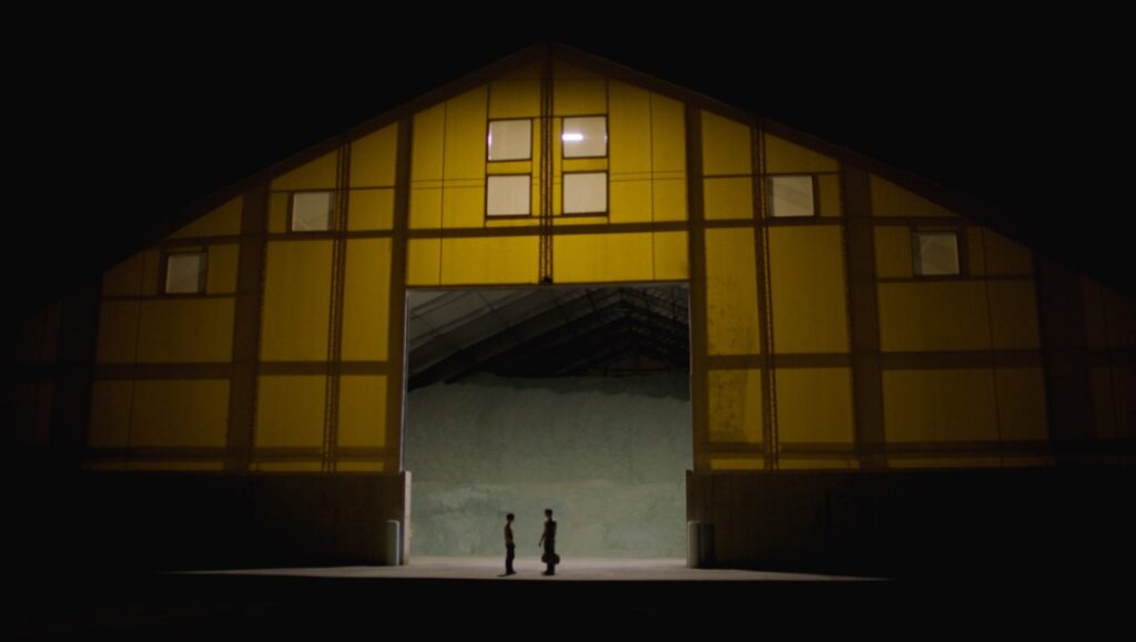 Stay Awake — Jamie Sisley Stay Awake film still: Two figures stand before a large, yellow warehouse-like structure at night.