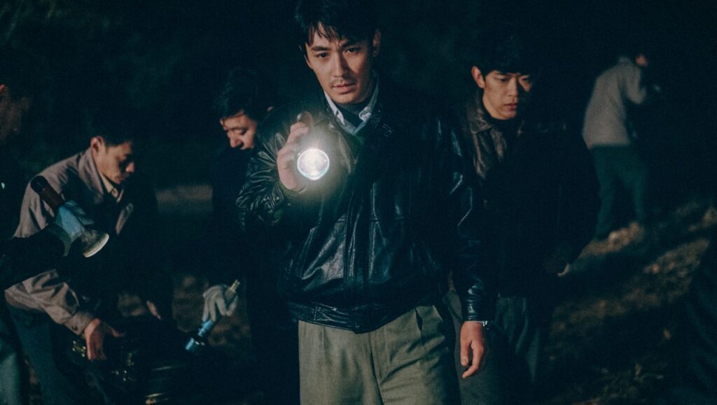 Only the River Flows film scene. A man holds a flashlight in a dark outdoor setting with other men.