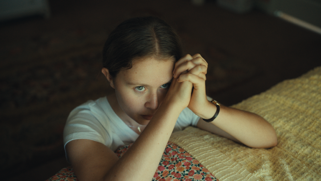 The Starling Girl movie scene: Elsie Fisher as a young woman with hands clasped in prayer. Emotional drama film.