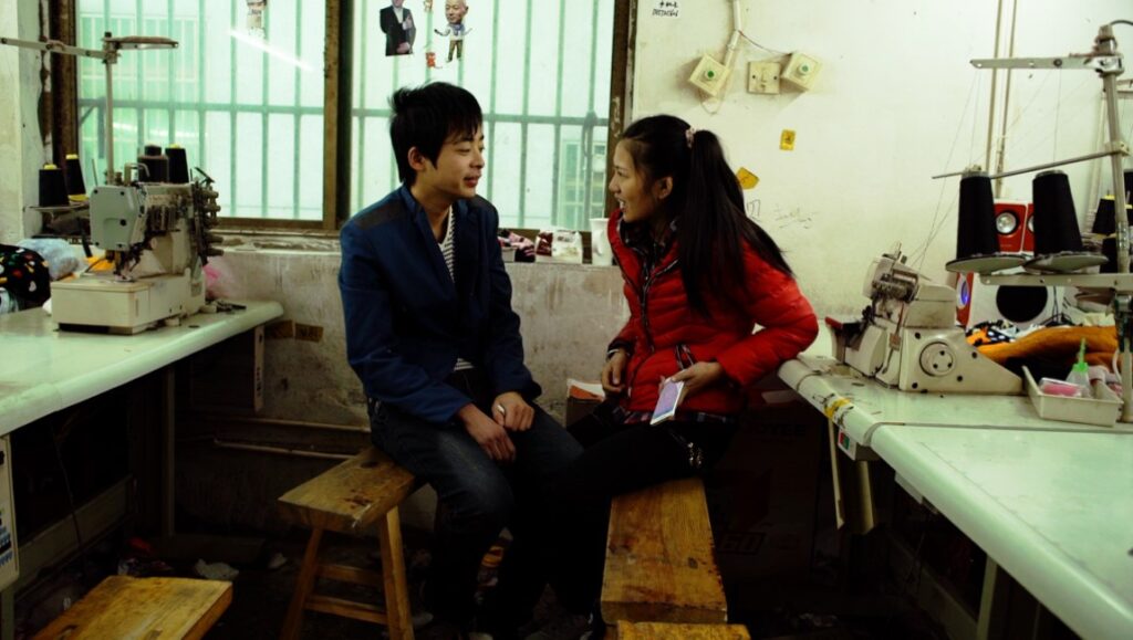 Wang Bing's Youth (Spring): Two young workers chat in a Chinese textile factory. Sewing machines and spools of thread visible.