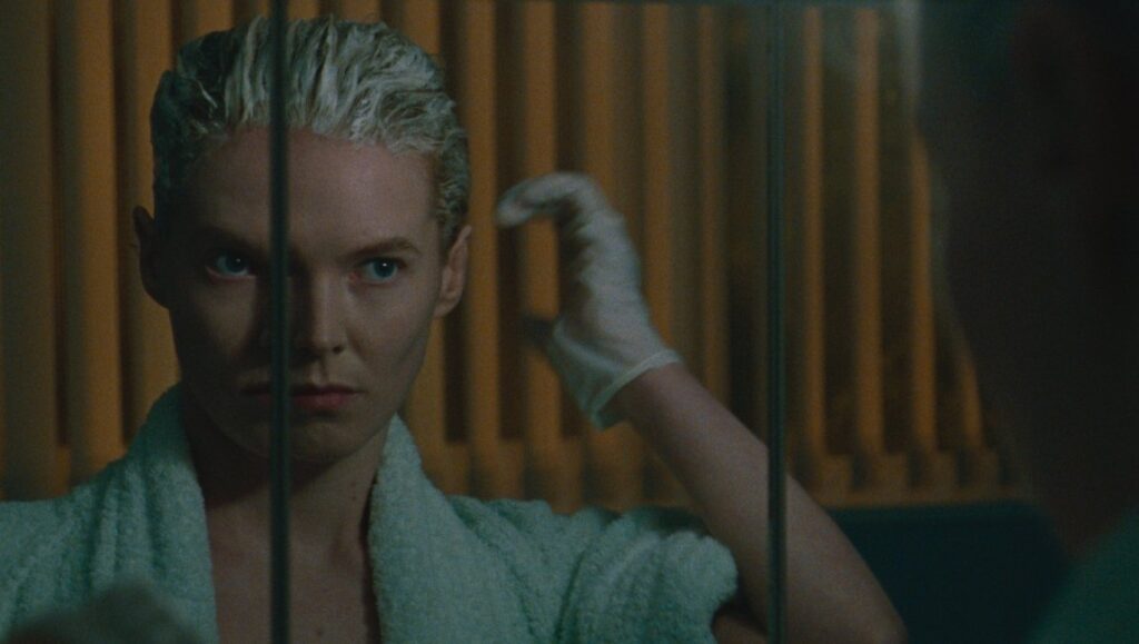 Blue Jean movie still: Woman in robe with dyed hair, wearing gloves, looks in mirror. Georgia Oakley film.
