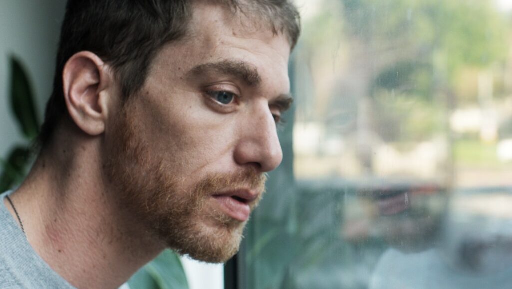Idan Haguel in Concerned Citizen film still. Close-up of a man with a beard looking out a window. Guy Sahaf photography.