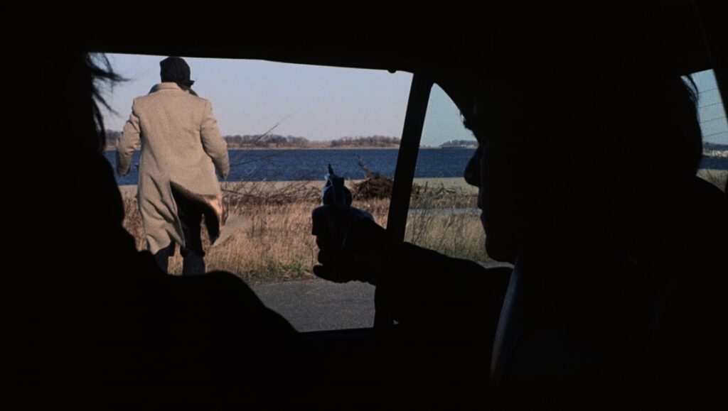 Scene from The Friends of Eddie Coyle movie. Man with gun in car, another man walking away toward water. Crime, film.