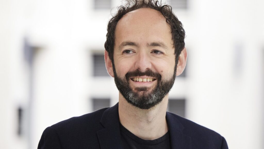 Portrait of Julien Rejl, smiling man with a beard, wearing a black suit jacket.