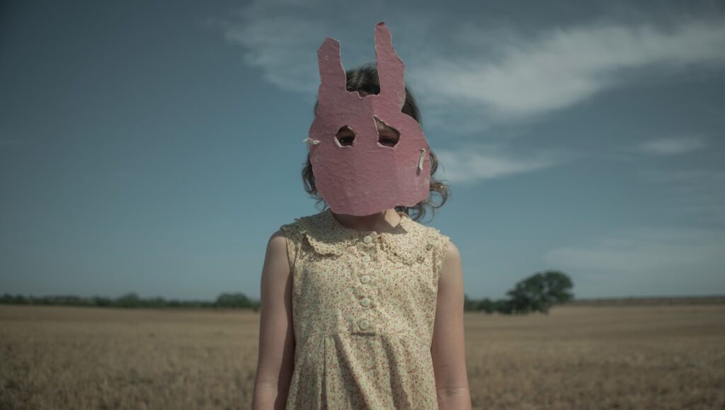 Run Rabbit Run Netflix film: Girl in dress wearing a pink rabbit mask in a field. Sarah Snook movie.