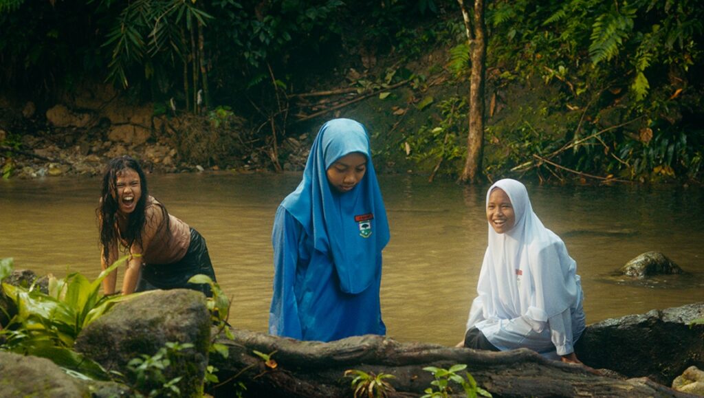 Tiger Stripes film still. Three girls in hijabs stand in a river. One screams. Jungle backdrop.