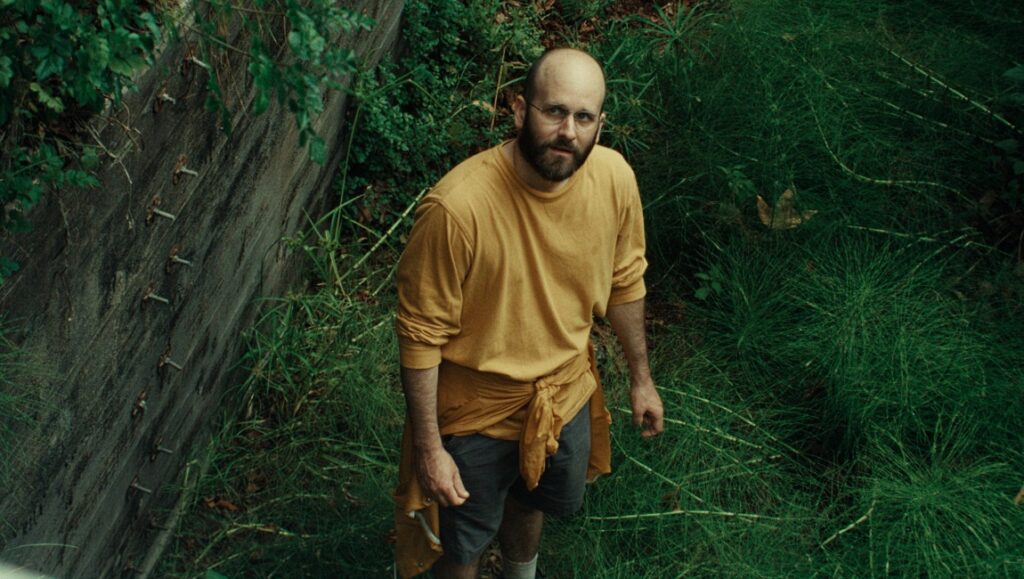 Al Warren, Dogleg promotional image. Bald man with beard in yellow shirt, standing near a concrete wall and green plants.